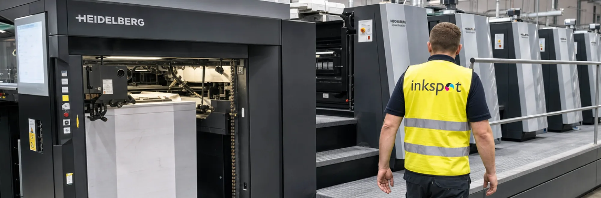 An operator walking the length of a multi-colour Heidelberg offset press, with a pallet of sheets loaded at the feeder.
