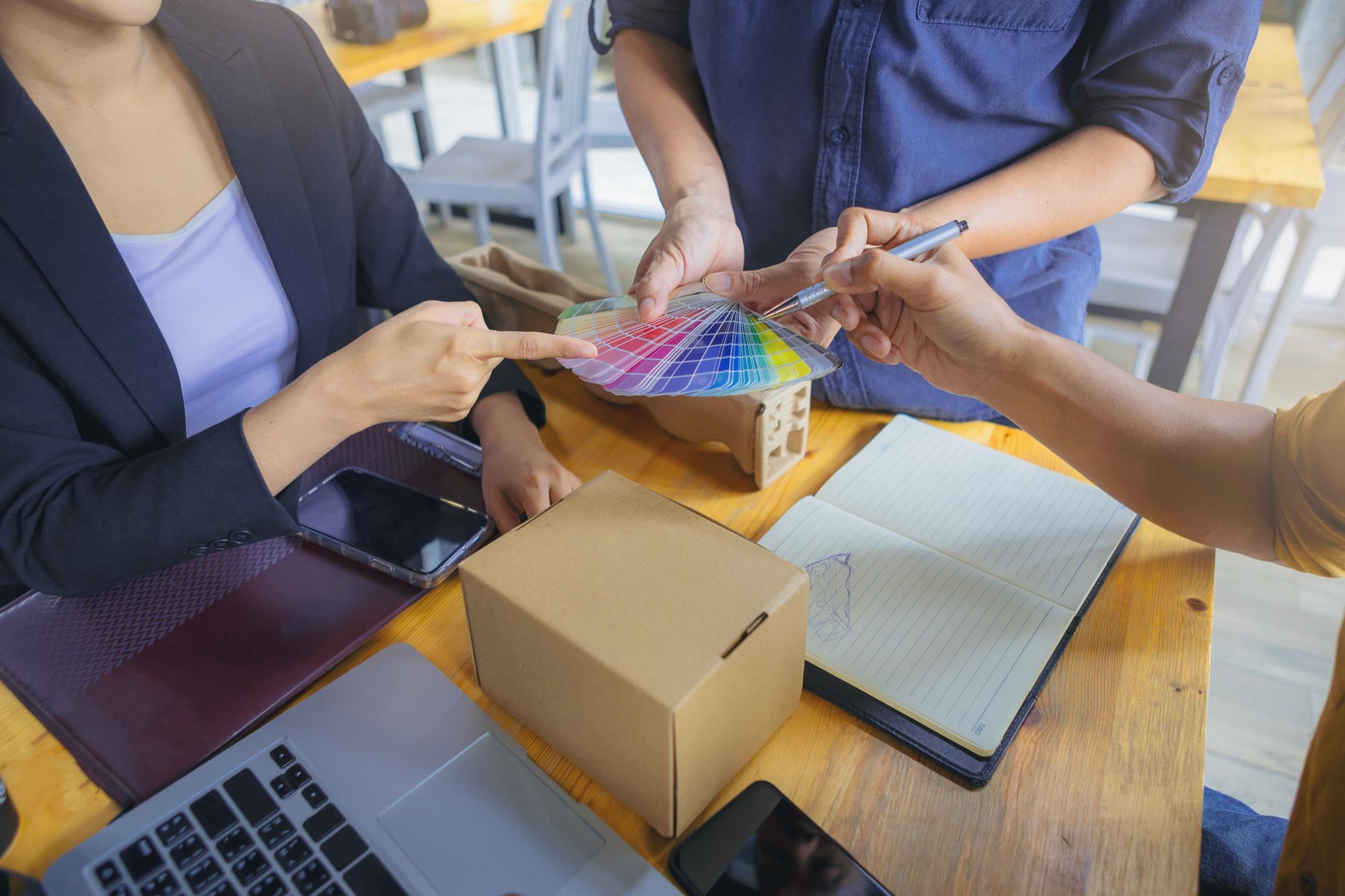 Two people comparing a Pantone swatch fan over a kraft carton sample, with a notebook and sketches on the table.