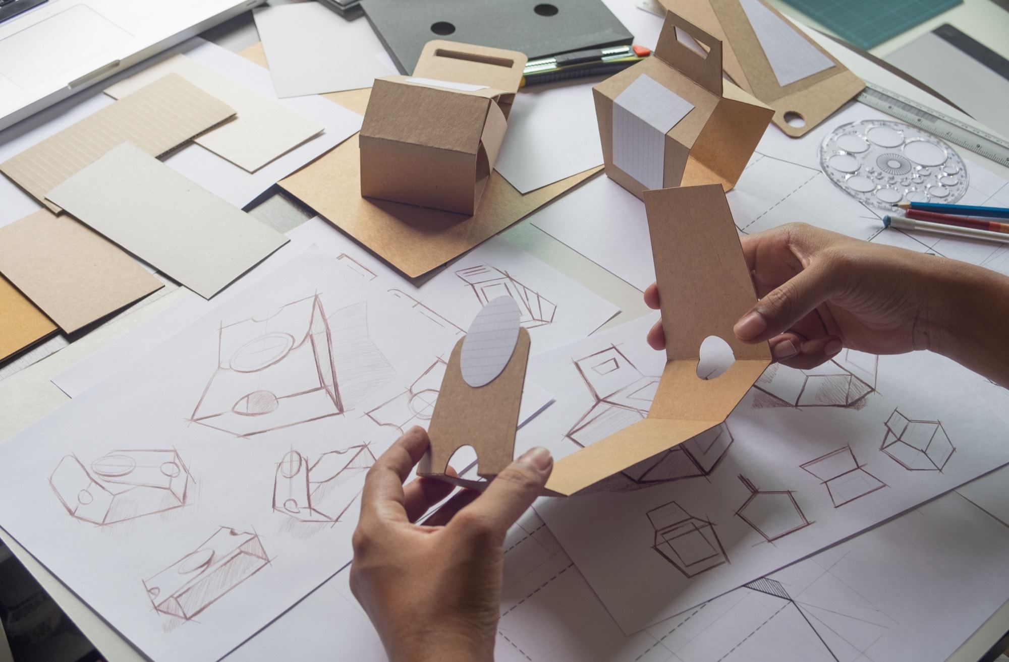 A packaging designer holding folded kraft carton samples over pencil structural sketches and dieline drawings.