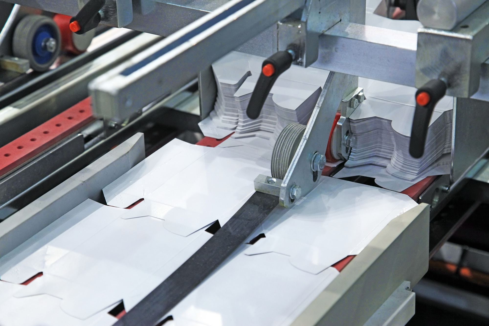 Die-cut white cartons moving through a folder gluer, with stacked blanks waiting behind.