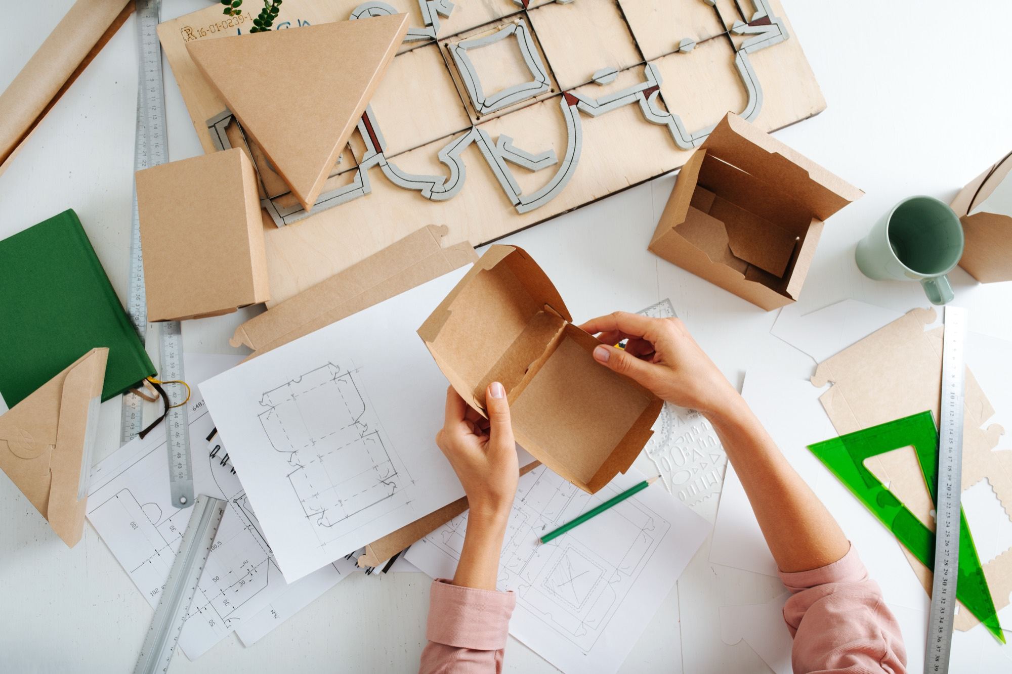 Overhead view of folded kraft carton prototypes, dieline drawings and a wooden cutting forme on a studio bench.